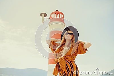 A girl in a yellow-orange suit on the beach against the background of the sea, mountains and a lighthouse. Fashionable woman in Stock Photo