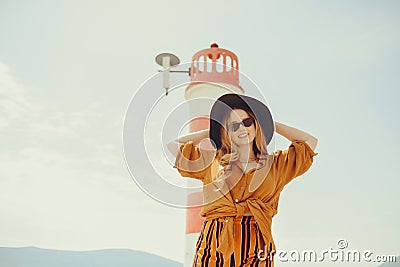 A girl in a yellow-orange suit on the beach against the background of the sea, mountains and a lighthouse. Fashionable woman in Stock Photo