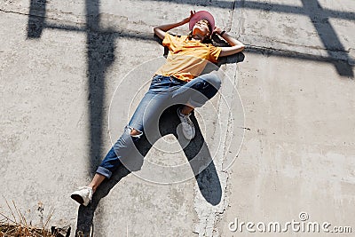 The girl in yellow t-shirt, jeans and pink visor is laying on a concrete slab on a sunny day outside Stock Photo