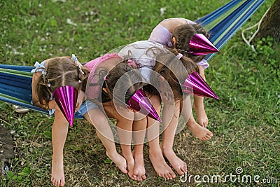 Girls in festive hats are sitting on hammock, hiding faces joking on birthday Stock Photo