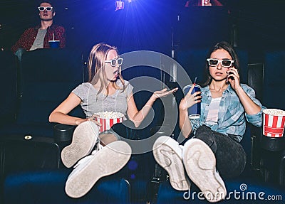 Girls are sitting in chairs in cinema hall. Brunette is talking on the phone while her friend is making remark to her Stock Photo