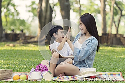 Giving high-five, mother and son holding a notebook and sitting picnic in the park Stock Photo