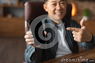 Glad adult asian man pointing finger at smartphone with empty screen at table in home office interior Stock Photo