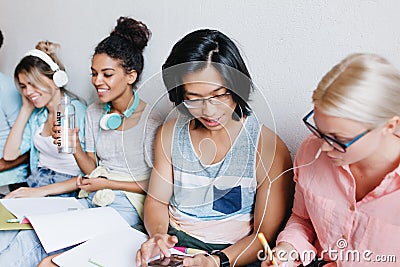 Glad asian student typing message on smartphone while his charming blonde friend writing lecture. Indoor group portrait Stock Photo