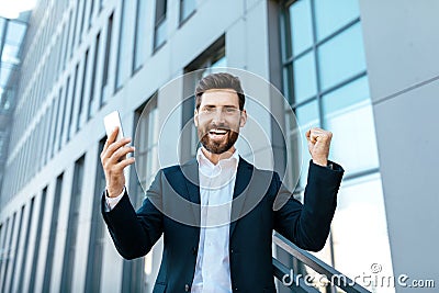 Glad caucasian millennial man with beard in suit with smartphone rejoices to victory and raises hands up Stock Photo