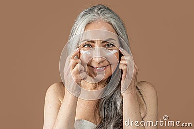 Glad elderly caucasian female with natural beauty applies cream on her face, fight with wrinkles Stock Photo