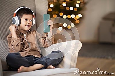 Glad european small kid in wireless headphones calls by phone, sits in armchair in living room interior Stock Photo