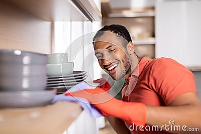 Glad millennial black man in rubber gloves wipes dust from shelf with dishes enjoy purely in white kitchen Stock Photo