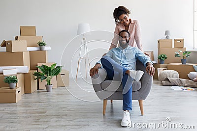 Glad smiling young afro american husband sitting in armchair, wife standing near him in apartment with cardboard boxes Stock Photo