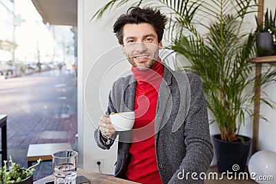 Glad young attractive unshaved brunette guy with trendy haircut holding cup of tea in raised hand and looking positively at camera Stock Photo