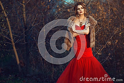 Glam model with updo hair and beautiful make up wearing posh red fishtail dress and luxurious mink vest standing in dry bushes Stock Photo