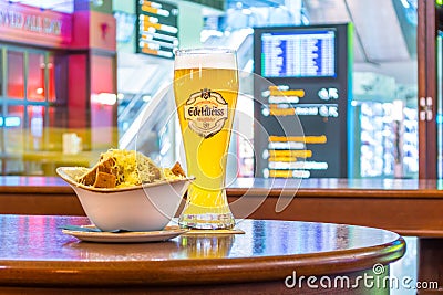 A glass of beer Edelweiss and a plate of breadcrumbs and cheese. In the background, online board displays flights to the airport. Editorial Stock Photo