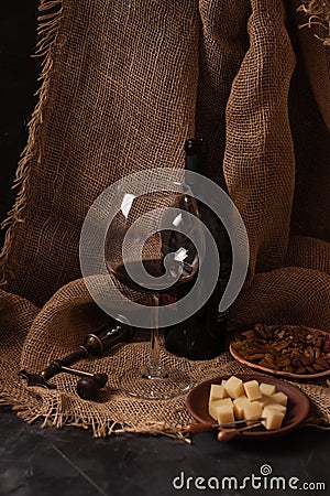 Glass and bottle of red wine with cheese, raisins, and nuts on sackcloth, dark background Stock Photo