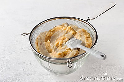 A glass bowl with pureed quince in a sieve and a silicone spatula for rubbing on a light gray background.Stage of Stock Photo