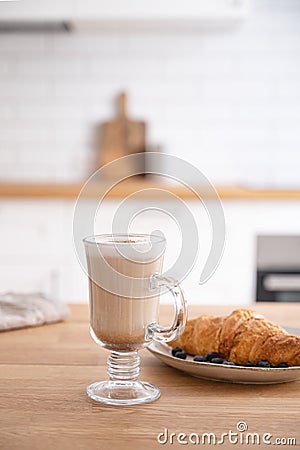 Glass of fragrant cappuccino coffee with cinnamon and croissant on a wooden table against the background of a white kitchen in th Stock Photo