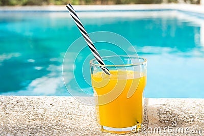 Glass of freshly pressed tropical fruits orange juice with striped straw standing on deck of swimming pool. Bright sunlight Stock Photo