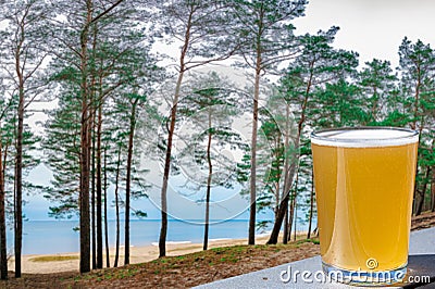 Glass of light beer against view of coniferous forest with pine trees and blue sea. Baltic sea coast Stock Photo