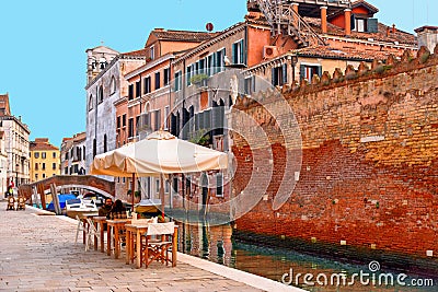 Glimpse of Venice with one of its canals with boats, historic buildings and people drink and relax in outdoor table and chairs of Editorial Stock Photo