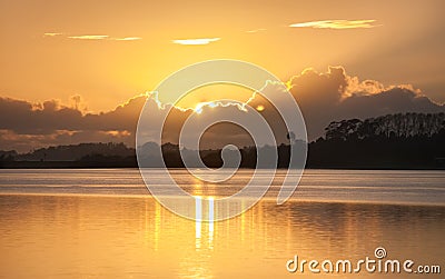 Glow of rising sun behind clouds across bay. Stock Photo