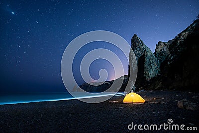 Glowing camping tent on a beautiful sea shore with rocks at night under a starry sky Stock Photo