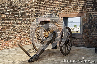 A gold and brown cannon surrounded by the red brick walls of a military fort on a brown wooden floor at Old Fort Jackson in Stock Photo