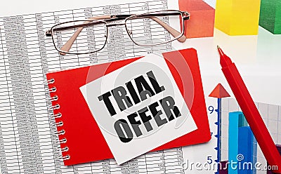 Gold-rimmed glasses, a red pen, color tables and a red notebook with a white card with the text TRIAL OFFER on the desktop. Stock Photo