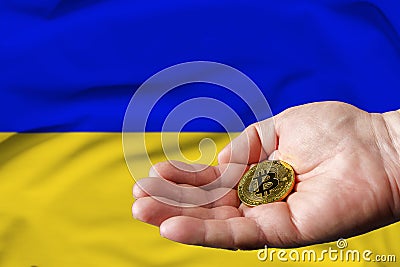 Golden bitcoin coin in man`s hand, Ukrainian flag in the background Stock Photo
