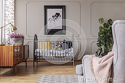 Golden lamp and heather in patterned pot on the vintage cabinet in grey baby room interior with crib and armchair Stock Photo