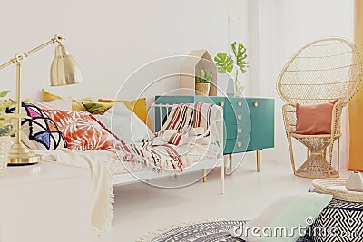 Golden lamp on nightstand in boho girl`s bedroom with colorful bedding on bed, green wooden cabinet and peacock chair with pillow Stock Photo