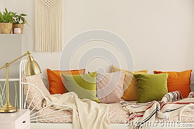 Golden lamp on the nightstand next to olive green, pastel pink, yellow and orange pillows on single metal bed with patterned Stock Photo