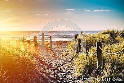 Golden sunset over sandy pathway with grass reeds and wooden posts on each side leading to a beautiful sea bay Stock Photo