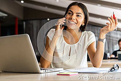Good business talk. Cheerful young beautiful woman talking on mobile phone and using laptop with smile while sitting at Stock Photo