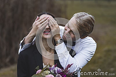 Good-humored young attractive Scandinavian groom in elegant white shirt covering the eyes of his newlywed Turkish wife Stock Photo