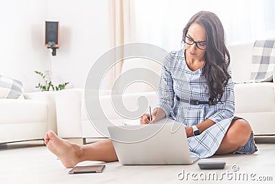 Good looking brunette in a dress sits barefoot on her living room floor, working on a notebook, writing notes Stock Photo