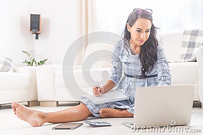 Good looking brunette in a dress sits barefoot on her living room floor, working on a notebook, writing notes Stock Photo