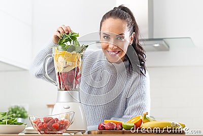Good looking girl piles colorful mix of fruit and green leaves into a blender at home in the kitchen Stock Photo