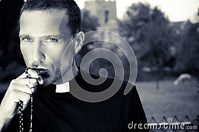 Good looking priest kisses his rosary with church, field and alpaca in background Stock Photo