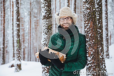 Good looking smiling male wears warm winter clothes holds firewood, stands near tree, spends free time with friends in winter fore Stock Photo