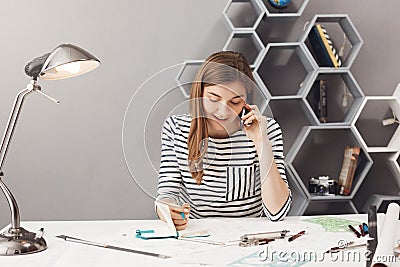 Good-looking young entrepreneur designer with dark hair in striped shirt talking on phone with customer discussing Stock Photo