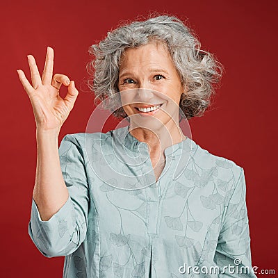 Good, perfect and okay with a senior woman hand gesture or sign in support or positive backing in studio against a red Stock Photo