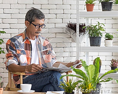 A goold looking senior Asian man wearing eyesglasses sitting and drink coffee and reading the newspaper with happiness. Idea for Stock Photo