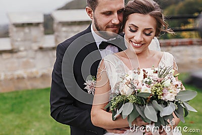 Gorgeous bride with modern bouquet and stylish groom gently hugging and smiling outdoors. Sensual wedding couple embracing. Roman Stock Photo