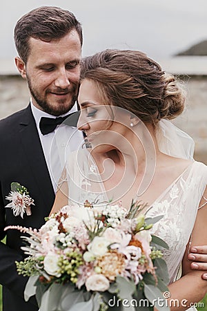 Gorgeous bride with modern bouquet and stylish groom gently hugging and smiling outdoors. Sensual wedding couple embracing. Roman Stock Photo