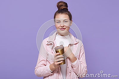 Gorgeous charming young female with hair knot holding large thermo mug, enjoying freshly made coffee, wearing white shirt and Stock Photo