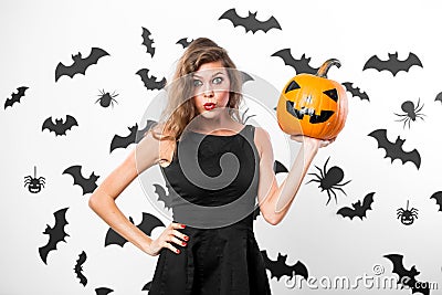 Gorgeous girl wearing black dress holds horror halloween pumpkins on the background of the wall with bats Stock Photo