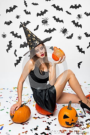 Gorgeous girl wearing black dress, witch hat and high heels sits on the pumpkin and holds Halloween pumpkin in her hand Stock Photo