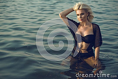 Gorgeous glam tanned blond woman wearing trendy black two piece strapless swimsuit and summer tunic sitting in the transparent sea Stock Photo