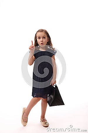 Gorgeous pretty 4 years old little Caucasian European girl wearing evening attire, hold a shopping black packet and point on a Stock Photo