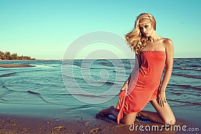 Gorgeous slim blond model in coral red strapless dress standing on knees in the sea water and looking aside Stock Photo