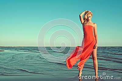 Gorgeous slim blond model in red strapless dress with flying train standing on tiptoe in the sea water Stock Photo